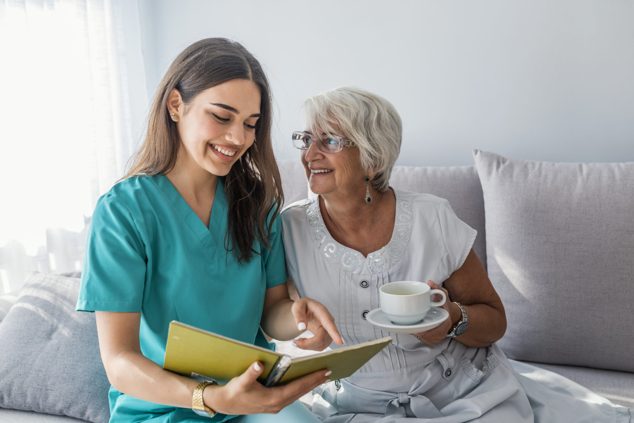 Happy patient is holding caregiver for a hand while spending time together. Elderly woman in nursing home and nurse. Aged elegant woman and tea time at nursing home